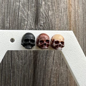 Puede incluir: Tres pequeños pendientes de calavera en negro, rojo y beige. Los pendientes se muestran sobre una superficie blanca.