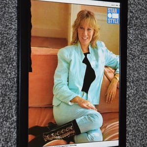 Op de afbeelding: Ingelijste magazinecover met een vrouw in een lichtblauw pak en een zwarte top. De afbeelding heeft de tekst "STAR STYLE" in een blauwe doos. De vrouw draagt zwarte laarzen met een decoratief patroon.
