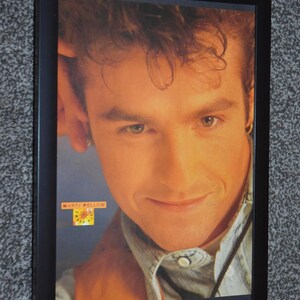 Puede incluir: Un retrato enmarcado de Marti Pellow que muestra un primer plano de un rostro con cabello castaño, ojos claros y una sonrisa sutil. El sujeto viste una camisa de mezclilla azul claro y una prenda marrón. El texto MARTI PELLOW aparece en un recuadro amarillo.