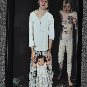 Könnte beinhalten: Ein gerahmtes Foto von Kurt Cobain, Courtney Love und Frances Bean in ihrem Zuhause im Jahr 1993. Das Bild zeigt drei Personen in Freizeitkleidung, mit Text in der oberen linken Ecke, der die Personen und den Ort der Szene beschreibt.