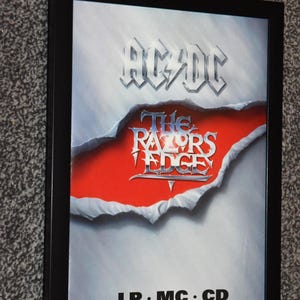 May include: Framed poster for the AC/DC album "The Razors Edge." The album title is in a stylized font, with the band's name at the top. The background is silver with a torn paper effect revealing a red background.