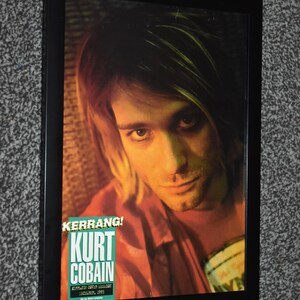 May include: Framed poster featuring Kurt Cobain with long blonde hair, wearing a red shirt. The poster includes the text "KERRANG!" and "KURT COBAIN" in a green and white font. The poster is in a black frame.