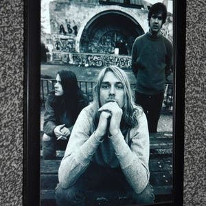 May include: A black and white framed photograph featuring three individuals sitting outdoors. The central figure has long hair and rests their chin on clasped hands. To the left, a person holds a cigarette, and to the right, a person stands.