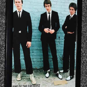 Op de afbeelding: Een ingelijste foto van de band The Jam in 1977, met drie personen in zwarte pakken en dassen voor een lichtblauwe bakstenen muur. De tekst luidt: The Jam in 1977: from left Rick Buckler, Paul Weller and Bruce Foxton. ONE OF THE GREATEST BANDS AT THEIR VERY BEST.