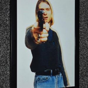 May include: Framed photograph featuring a person with long blonde hair pointing a handgun directly at the viewer. The person is wearing a dark sweater, blue jeans, and a black belt. The image is set against a white background.
