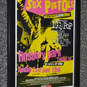 May include: A framed concert poster for the Sex Pistols Filthy Lucre tour at Finsbury Park on Sunday 23rd June 1996. The poster features a high-contrast yellow and black graphic with pink text, listing Iggy Pop and other special guests.