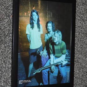 May include: A framed rectangular poster featuring a photograph of the band Nirvana with three members. The image shows a yellow t-shirt, a green striped shirt, and an electric guitar. Text at the bottom reads: NIRVANA: Smells Like Teen Spirit... and causing a stink in Kerrang!