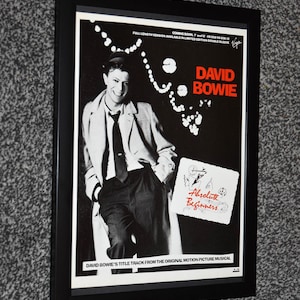Könnte beinhalten: Schwarz gerahmtes Poster mit einem Schwarz-Weiß-Bild von David Bowie. Der Titel "David Bowie" ist rot. Das Poster enthält den Text "Absolute Beginners" und weitere Informationen. Das Poster ist in einem schwarzen Rahmen.