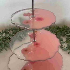Op de afbeelding: Een serveerschaal met drie niveaus met roze en witte bloemmotieven. De randen van elk niveau hebben een geschulpte vormgeving. De schaal wordt ondersteund door een zilveren metalen staaf. De schaal is versierd met roze bloemen en glitter.