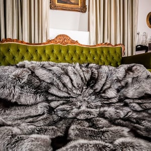 Op de afbeelding: Een luxe, zilver-grijze imitatiebont deken over een groene fluwelen bank. De deken heeft een dichte, getextureerde uitstraling, waardoor een zachte en uitnodigende uitstraling ontstaat. De achtergrond omvat een ingelijst schilderij en elegante gordijnen.