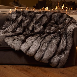 Puede incluir: Una lujosa manta de piel sintética en tonos grises y negros está sobre un sofá marrón oscuro. La manta de felpa tiene un aspecto texturizado, creando una estética acogedora y elegante. Un accesorio decorativo para el hogar.