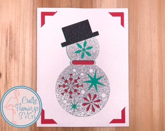 Puede incluir: Una tarjeta blanca con un borde rojo presenta un muñeco de nieve de purpurina plateada con un sombrero de copa negro, diseños de copos de nieve verdes y rojos y una bufanda roja. La tarjeta está sobre un fondo de madera.