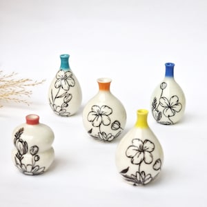 May include: Five small ceramic vases with a white background and black floral designs. The vases have different coloured rims: blue, yellow, orange, and teal.