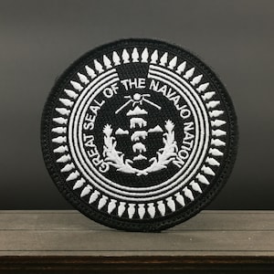 May include: A circular black and white embroidered patch featuring the Great Seal of the Navajo Nation. The design includes a central stack of animals, mountains, and corn stalks, surrounded by a border of arrowheads and the text Great Seal of the Navajo Nation.