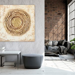 Puede incluir: Una pieza de arte mural cuadrada con un motivo circular central compuesto por capas de pintura dorada texturizada. El fondo consiste en pintura espesa de color crema y blanco, creando una superficie elevada sobre el lienzo.