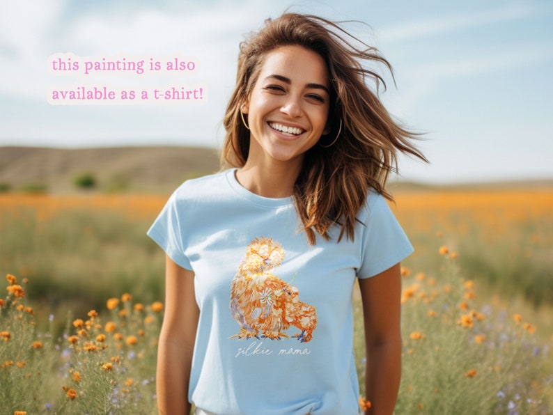 A smiling young woman in a wildflower farm field is wearing a light blue t-shirt printed with silkie chickens. Printed below the painted is the text Silkie Mama in a handwritten font. The text says that This painting is also available as a t-shirt.