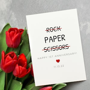 May include: A white card with the words "ROCK PAPER SCISSORS" crossed out, with the words "HAPPY 1ST ANNIVERSARY!" and the date "11.13.22" below. A red heart is next to the date. A bouquet of red tulips is in the background.