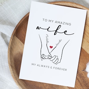 May include: A white card with black text that reads "To my amazing wife" with a stylized drawing of two hands holding each other with the names "Michelle" and "Amanda" written on the arms. The text "My always & forever" is at the bottom of the card.