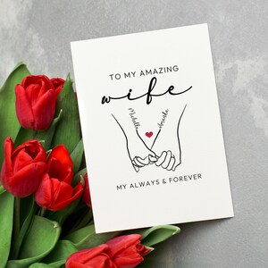 May include: A white card with black text that reads "To my amazing wife" with a stylized drawing of two hands holding each other with a heart in the middle. The names "Michelle" and "Brandon" are written on the hands. The text "My always & forever" is below the hands.