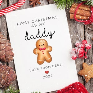 May include: A Christmas card with the text "FIRST CHRISTMAS AS MY daddy" and "LOVE FROM BENJI" with a gingerbread man illustration and a red heart. The year "2022" is also printed on the card. The card is on a wooden surface with Christmas decorations.
