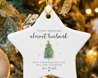 May include: White star-shaped Christmas ornament with gold string. The ornament has the text "TO MY AMAZING almost husband" and "NEXT CHRISTMAS WE'LL BE MR & MRS THOMPSON Love, Rachel" with a watercolor Christmas tree design.