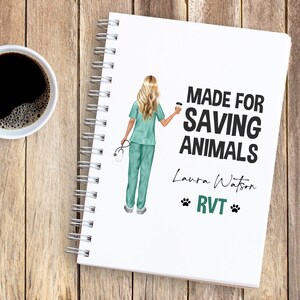 May include: A spiral-bound notebook with the text "MADE FOR SAVING ANIMALS" and the name "Laura Watson." The notebook features an illustration of a veterinary professional in scrubs. A cup of coffee sits next to the notebook on a wooden surface.