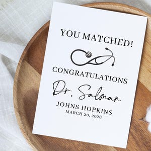 May include: A white card with the text "YOU MATCHED!" and "CONGRATULATIONS" in black. A stethoscope graphic is above the text. The card also includes the name "Dr. Salman", "JOHNS HOPKINS", and the date "MARCH 20, 2026".