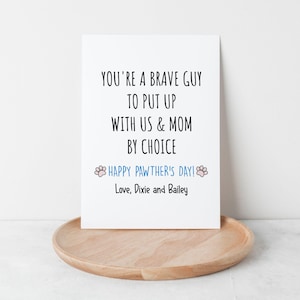 May include: A white greeting card with black text that reads "YOU'RE A BRAVE GUY TO PUT UP WITH US & MOM BY CHOICE." Below, in blue, it says "HAPPY PAWTHER'S DAY!" and "Love, Dixie and Bailey." The card rests on a wooden tray.