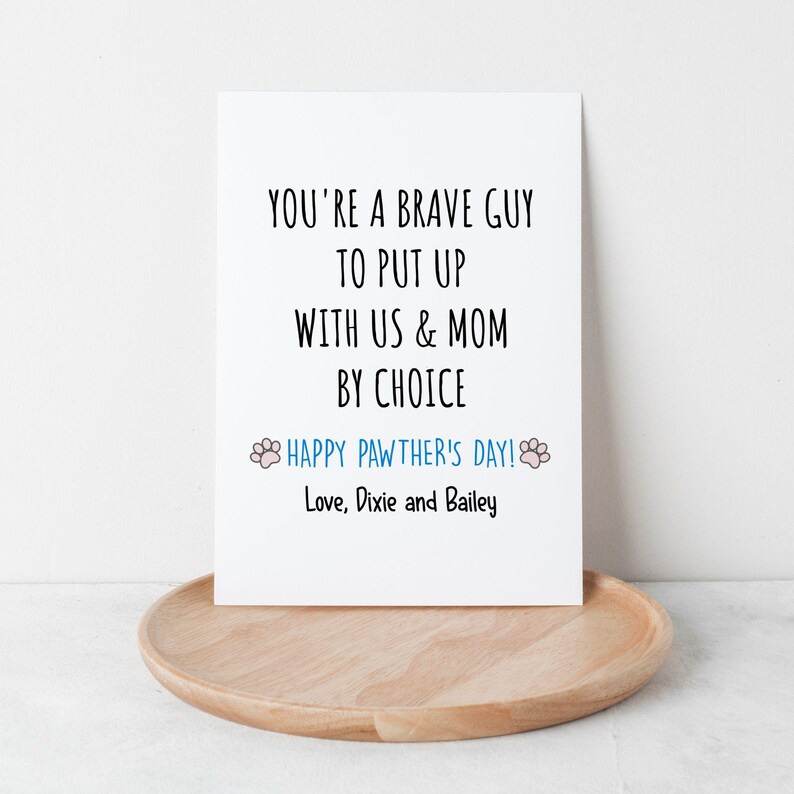 May include: A white greeting card with black text that reads "YOU'RE A BRAVE GUY TO PUT UP WITH US & MOM BY CHOICE." Below, in blue, it says "HAPPY PAWTHER'S DAY!" and "Love, Dixie and Bailey." The card rests on a wooden tray.