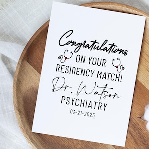 May include: White card with black text that reads "Congratulations on your residency match! Dr. Watson Psychiatry 03-21-2025". There are two red and black stethoscope drawings on the card.
