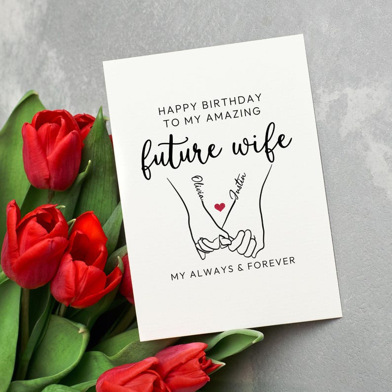 Może przedstawiać: Biała kartka z życzeniami z napisem "HAPPY BIRTHDAY TO MY AMAZING future wife" oraz imionami "Olivia" i "Justin" z czerwonym sercem. Na kartce znajduje się rysunek dw&oacute;ch trzymających się rąk. Czerwone tulipany w tle.