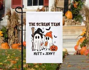 May include: A white Halloween garden flag featuring two cartoon ghosts. One ghost wears a witch hat and holds a broom, the other wears an orange beanie and holds a pumpkin spice drink. The flag reads "THE SCREAM TEAM" and "MATT & JENNY".