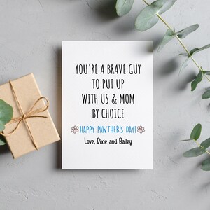 May include: A white greeting card with black text that reads "YOU'RE A BRAVE GUY TO PUT UP WITH US & MOM BY CHOICE." Below, in blue, it says "HAPPY PAWTHER'S DAY!" and "Love, Dixie and Bailey." A small gift box is on the left.