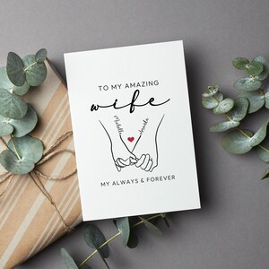 May include: A white card with black text that reads "To my amazing wife" with the names "Michelle" and "Amanda" written on the hands of a couple holding hands with a red heart in the middle. The text at the bottom reads "My always & forever".