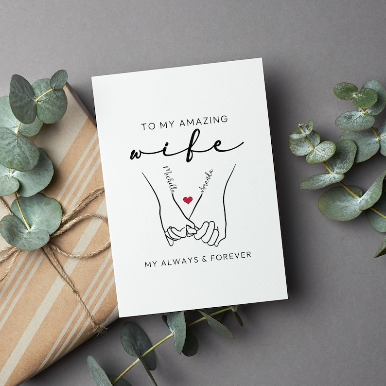 May include: A white card with black text that reads "To my amazing wife" with the names "Michelle" and "Amanda" written on the hands of a couple holding hands with a red heart in the middle. The text at the bottom reads "My always & forever".