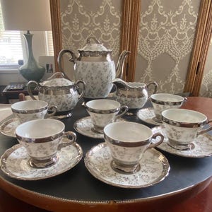 May include: A porcelain tea set featuring a white base with a delicate gray floral pattern and metallic silver trim. The set includes a teapot with a lid, a lidded sugar bowl, a creamer, and six teacups with matching saucers arranged on a table.