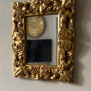 May include: A rectangular wall mirror featuring an ornate, thick gold-colored frame with detailed relief carvings of leaves, scrolls, and floral motifs. The reflective glass surface is clear and smooth, capturing a view of the surrounding room.