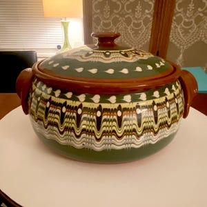 May include: A ceramic cooking pot with a lid. The pot is primarily green with a brown rim and handles. The pot features a decorative pattern of white, yellow, and black wavy lines and heart shapes. The lid has a matching pattern.