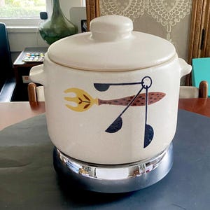 May include: Cream-colored ceramic casserole dish with a lid, featuring a retro design of a fork, spoon, and fish in yellow, brown, and black. The dish has side handles and sits on a chrome-colored base. A vintage kitchenware item.