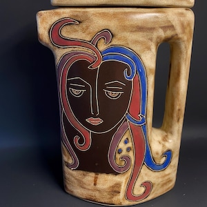 May include: A ceramic pitcher with a stylized face design. The face is painted in brown, red, blue, and purple. The pitcher has a lid and a handle.