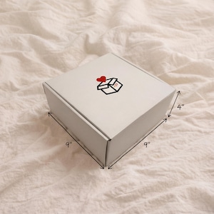 Closed white Caring Corner gift box on bedding with packed dimensions of 9 by 9 by 4 inches.