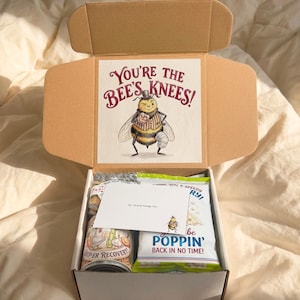 Close-up of the open knee replacement gift box with a personalized card, pun-labeled snacks, and “You’re the Bee’s Knees!” artwork inside the lid.
