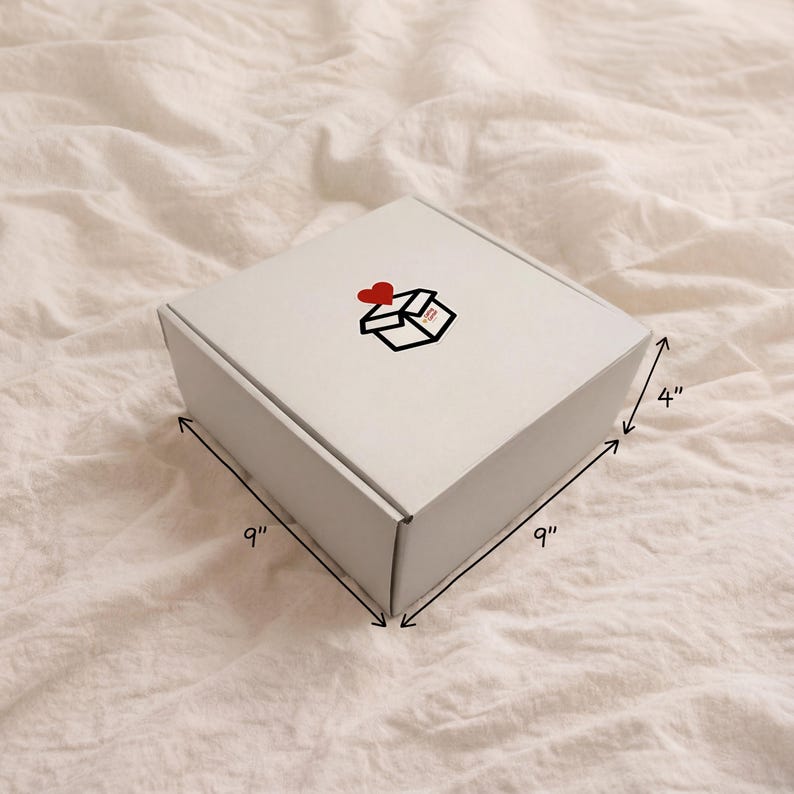 Exterior view of the closed white gift box packaging resting on white linens. Arrows indicate the box dimensions are 9 inches wide, 9 inches long, and 4 inches tall. Features a small "The Caring Corner" logo sticker on top.