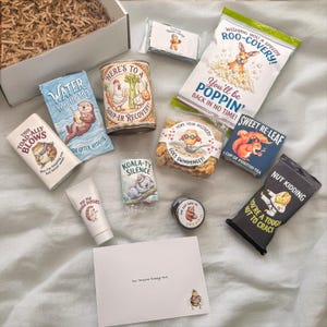 Flat lay of the knee surgery care package contents, including soup, popcorn, cookies, tea, honey, crackers, tissues, lotion, ear plugs, and a card.