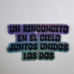 May include: A holographic sticker with the text "UN RINCONCITO EN EL CIELO JUNTOS UNIDOS LOS DOS" in black, with a glittery purple and blue gradient background. The sticker has a rounded, irregular shape.