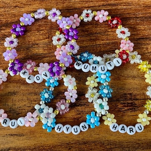 May include: Four beaded flower bracelets in various colors, including pink, purple, blue, and yellow. Each bracelet has white letter beads spelling out names or words: LOVE, RUI, HYUN, HARU, and WUMUTI. The bracelets are on a wooden surface.