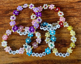 May include: Four beaded flower bracelets in various colors, including pink, purple, blue, and yellow. Each bracelet has white letter beads spelling out names or words: LOVE, RUI, HYUN, HARU, and WUMUTI. The bracelets are on a wooden surface.
