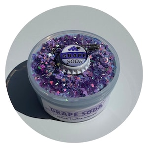 May include: A clear plastic container filled with purple slime, topped with purple heart, star, and flower-shaped glitter. A metal bottle cap charm labeled GRAPE SODA with a grape icon and a silver safety pin are placed on the surface of the slime.