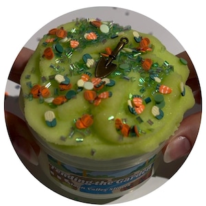 May include: A container of light green slime topped with small orange pumpkin, carrot, and corn-shaped sprinkles, green glitter, and a bronze-colored metal shovel charm. The label on the clear container reads Tending the Garden Moon Valley Slimes.