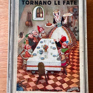 May include: A vintage hardcover book titled TORNANO LE FATE with a rectangular illustration on the cover. The scene depicts a woman in a white dress serving food to seven small figures seated around a white tablecloth on a checkered floor.